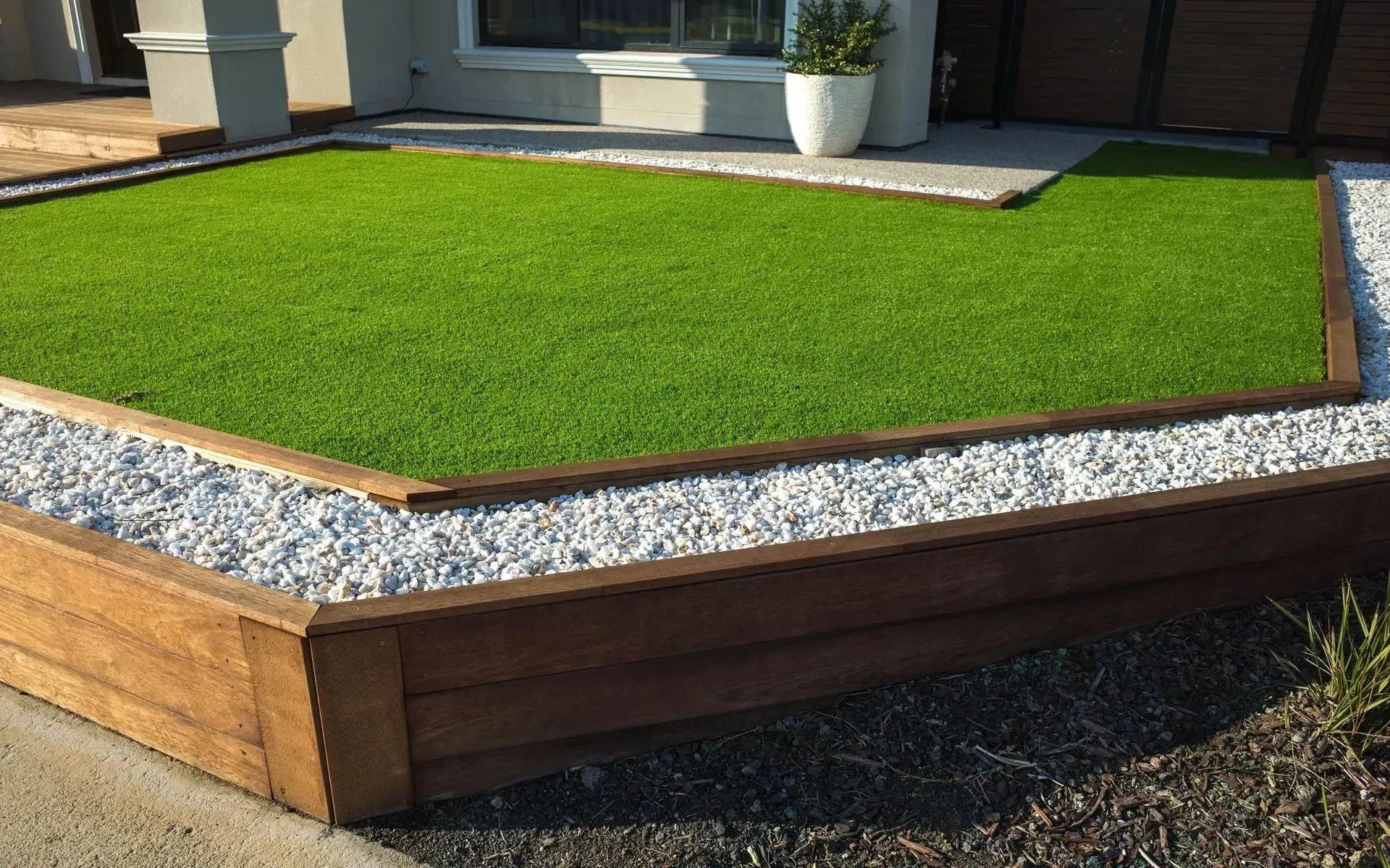 A raised lawn area featuring bright green artificial turf, bordered by white gravel, and enclosed by timber sleepers.