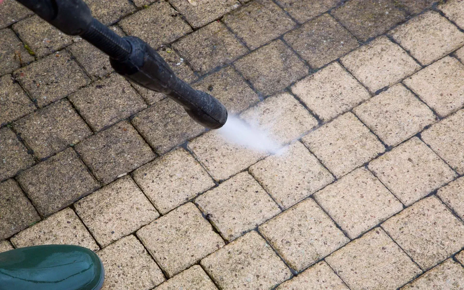 A pressure washer cleans a dirty light-colored brick patio, creating a visible path of clean stone.