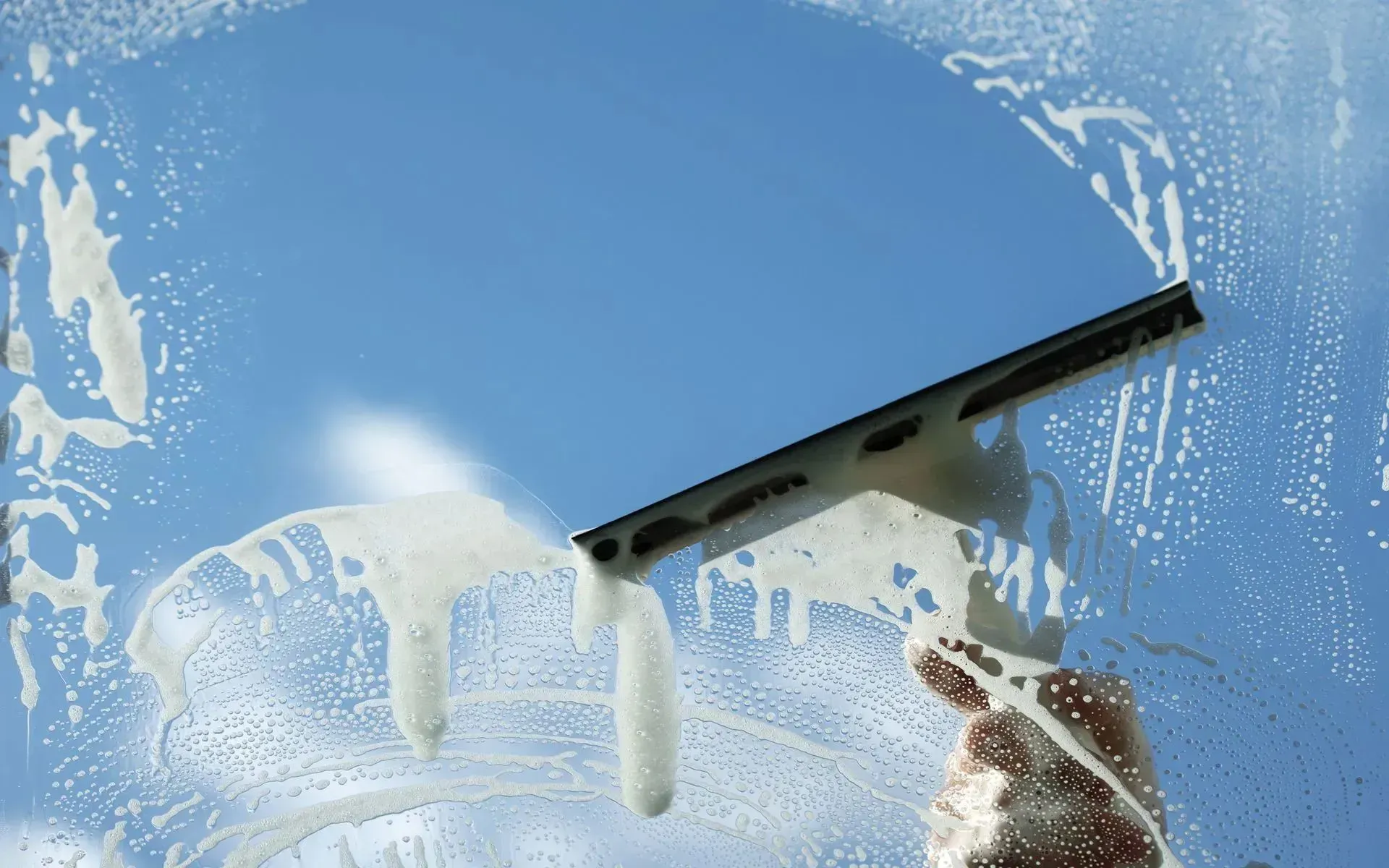 A hand uses a squeegee to wipe soapy water from a glass window against a clear blue sky.