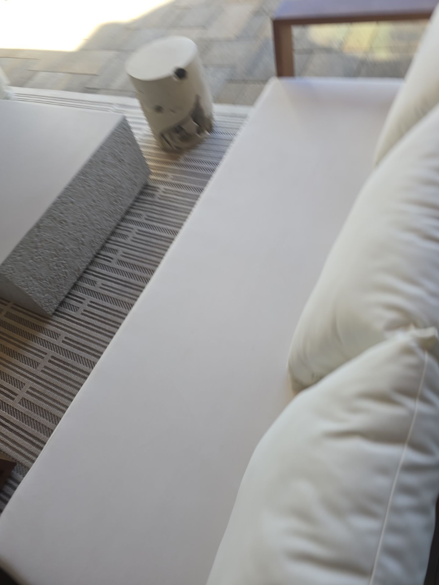 A white cushioned outdoor sofa sits next to a textured stone side table on a gray patterned rug.