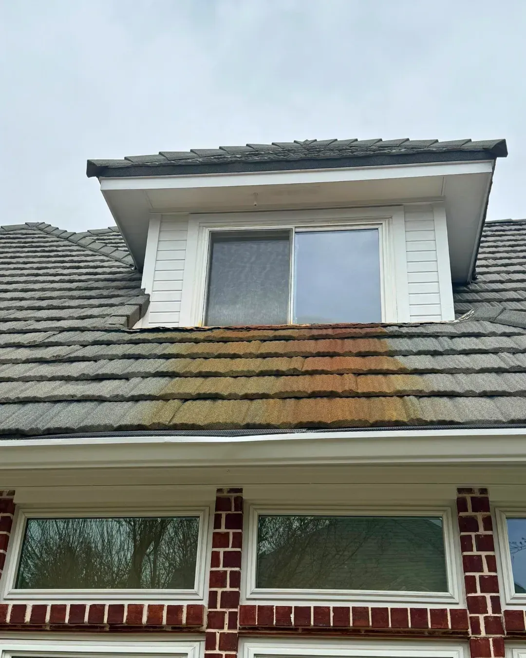 A dormer window on a gray tiled roof with a vertical streak of orange-brown rust staining the tiles below the window.