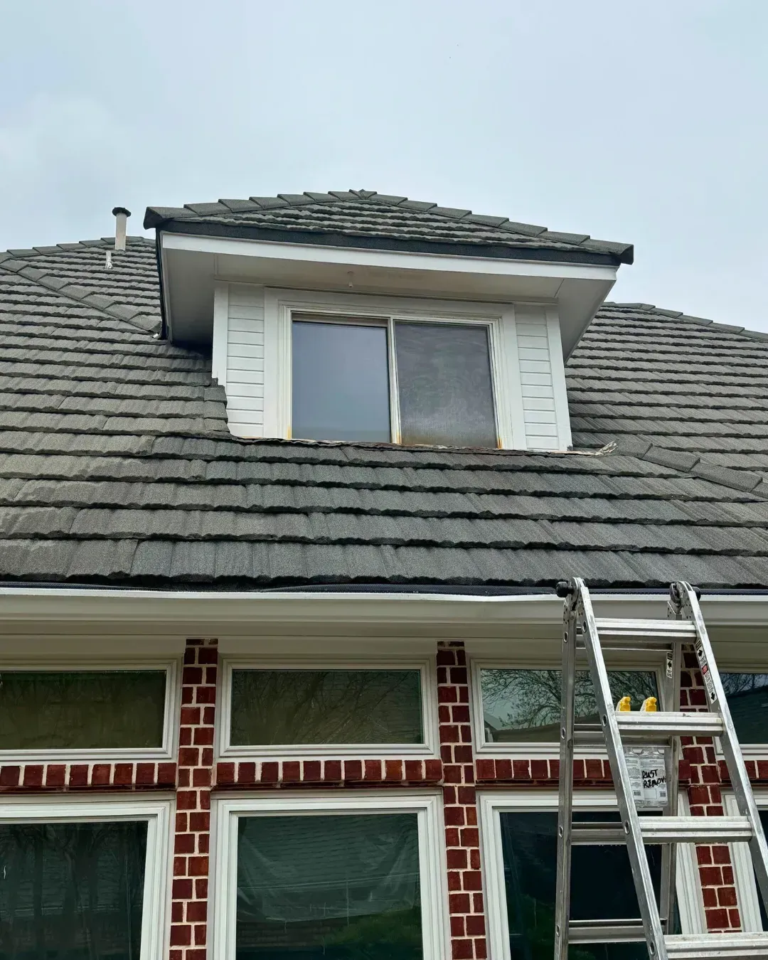 A dormer window with white siding and a dark tiled roof, set above a red brick wall with large glass windows and a ladder.