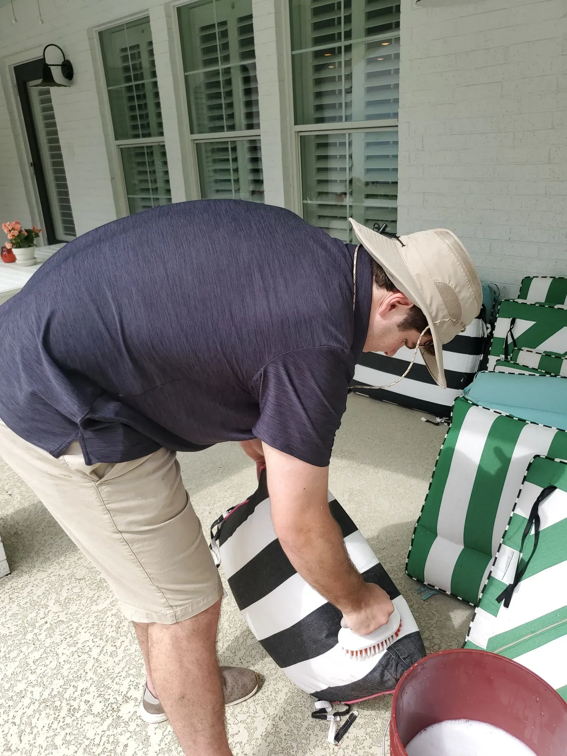 A person in a hat and shorts pours a white substance from a black-and-white striped bag into a red bucket on a patio.