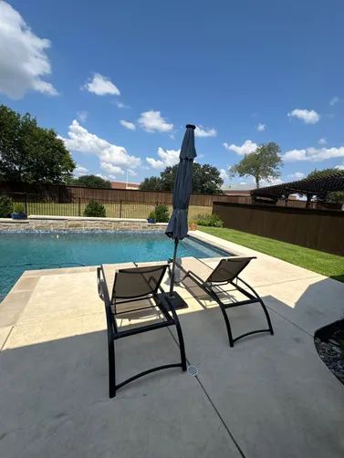 Two dark lounge chairs sit on a light-colored concrete pool deck next to a closed patio umbrella under a sunny blue sky.
