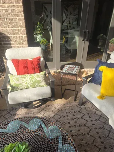 Two patio chairs with colorful pillows and a mosaic-topped table on a patterned rug in front of sliding glass doors.