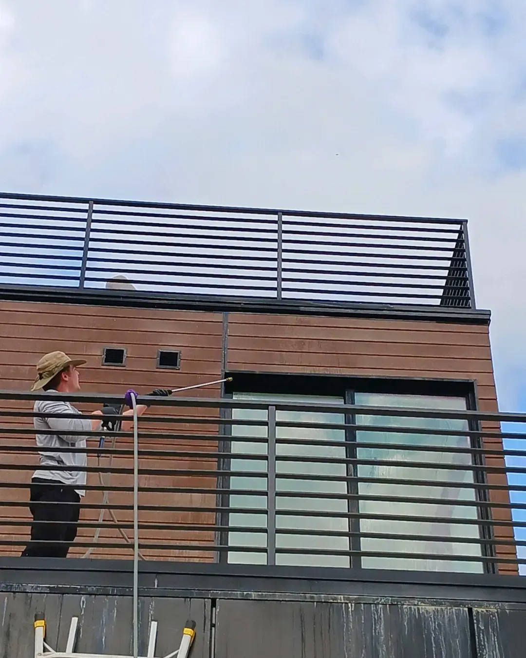 A worker in a hat uses a pressure washer to clean the exterior of a modern, brown building with a metal railing.