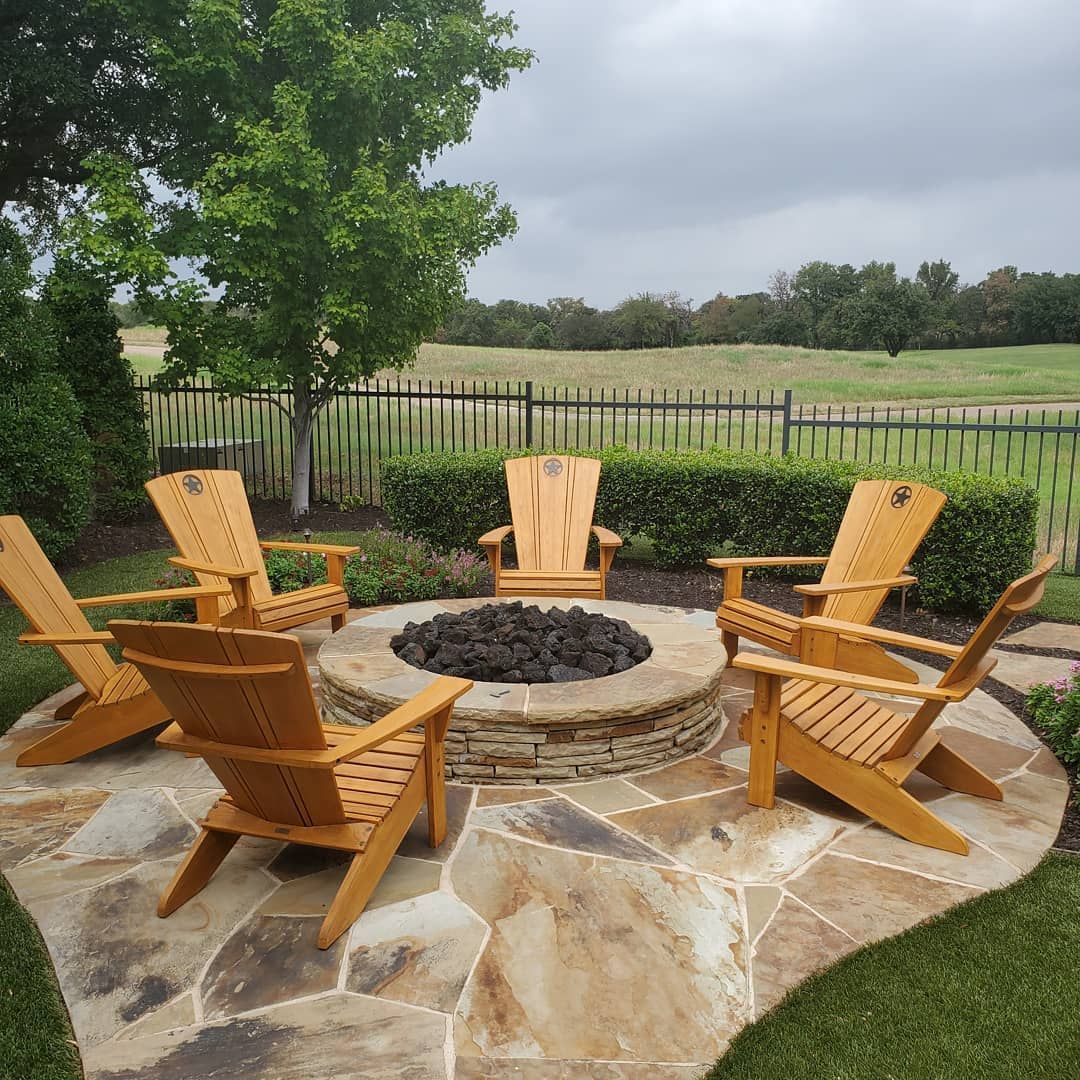 Five wooden Adirondack chairs arranged around a circular stone fire pit on a flagstone patio overlooking a grassy field.