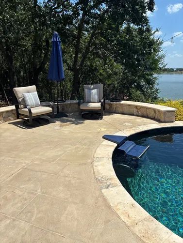 Two patio chairs and a blue umbrella on a stone deck beside a pool overlooking a lake with trees in the background.