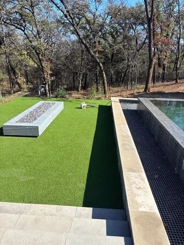 An outdoor patio area features a long rectangular fire pit, artificial grass, and a modern infinity pool with stone edges.