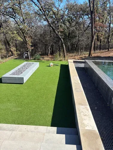 An outdoor lounge area with a rectangular fire pit on green turf next to a stone-edged swimming pool and wooded backdrop.