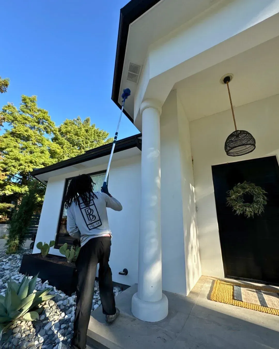 A person uses a long-reach pole tool to clean the exterior gutters of a white house with black trim and a black door.
