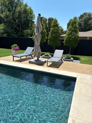 Two lounge chairs and a closed patio umbrella by a swimming pool, with a manicured lawn and privacy fence in the background.