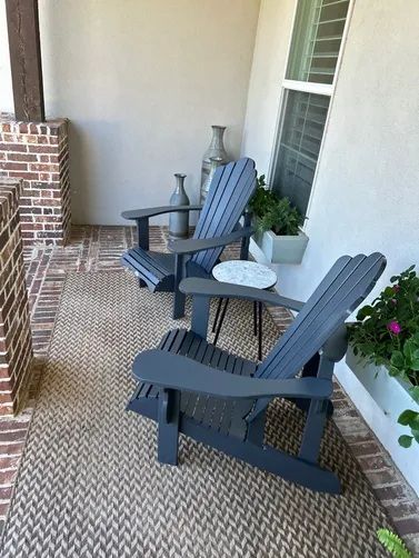 Two grey Adirondack chairs sit on a patterned rug on a covered porch with brick accents and a small potted plant.