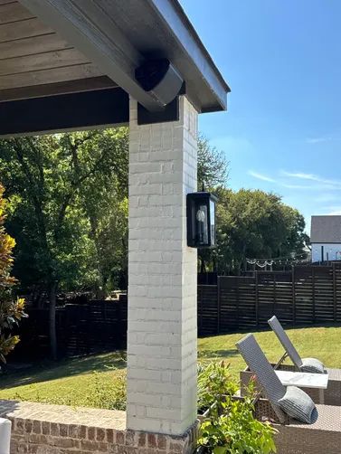 A painted white brick pillar supports a porch roof, featuring a modern black outdoor wall sconce and a mounted speaker.