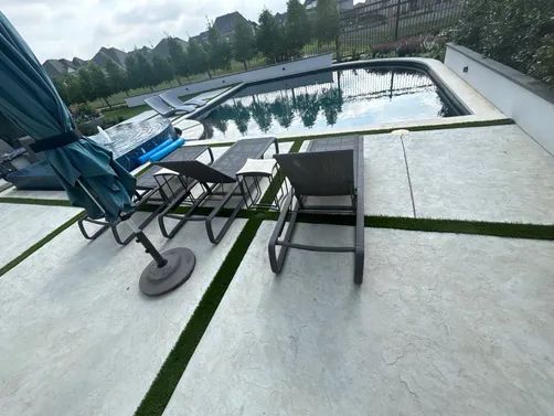 A patio with large stone pavers and artificial grass gaps, featuring lounge chairs near an outdoor swimming pool.