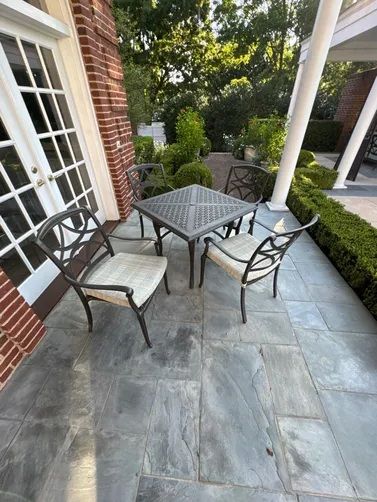 A patio with a square metal table and two chairs featuring light-colored cushions, set on gray stone tiles near a brick wall.
