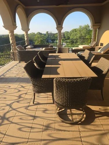 A patio with a large wooden dining table and wicker chairs, under an arched stone roof overlooking a green landscape.