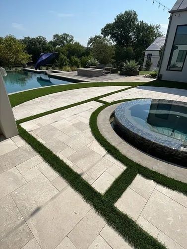 A modern patio with light stone pavers and grass inlays, featuring a circular hot tub, a swimming pool, and a fire pit.
