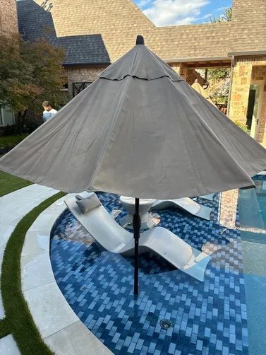 A tan patio umbrella stands over two white lounge chairs inside a tiled swimming pool at a backyard patio.