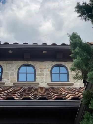 Two arched windows with dark frames centered on a stone wall above a curved, brown clay-tiled roof under a cloudy sky.