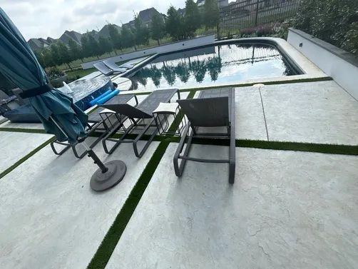 A modern backyard patio with gray pavers and turf borders, featuring two lounge chairs and a pool in the distance.