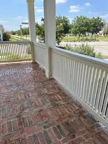 A covered front porch with a red brick floor and white wooden railings overlooking a neighborhood street on a sunny day.