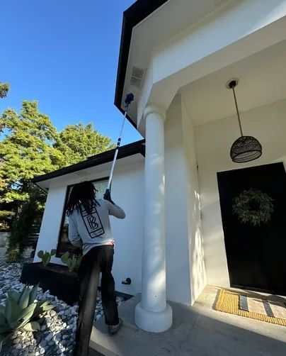 A person in a gray long-sleeved shirt uses a telescopic pole to clean the exterior roofline of a white house.