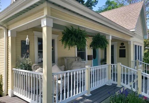 A yellow house with a white wrap-around porch, hanging ferns, outdoor wicker furniture, and a grey wooden deck.
