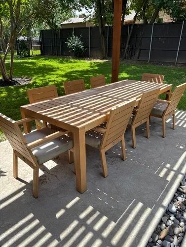 A long wooden dining table with eight matching chairs on a backyard patio with light-filtering shade.