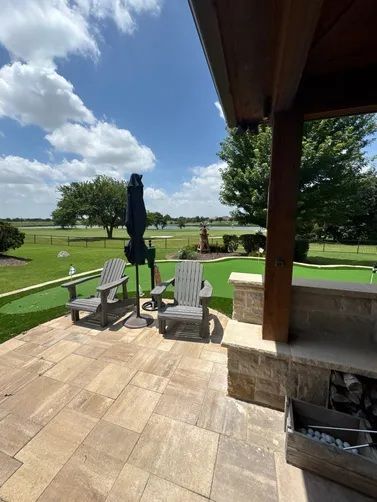 Patio with two grey adirondack chairs, a closed umbrella, and a putting green overlooking a lawn under a blue sky.
