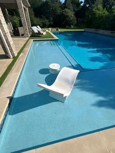 A white lounge chair and small table sit on the shallow sun shelf of a bright blue swimming pool.