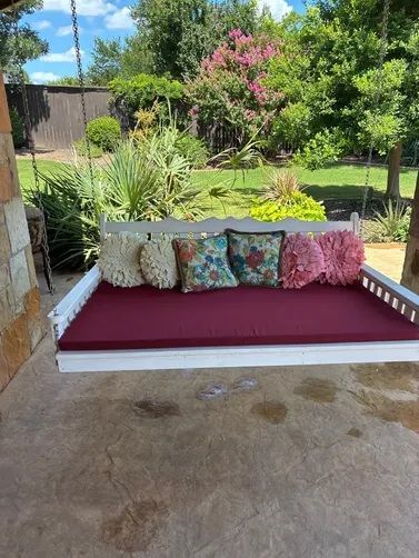 A white porch swing bed with a maroon cushion and various decorative pillows, situated on a patio overlooking a yard.