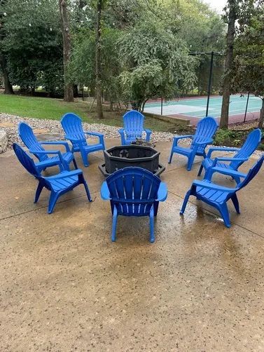 A circle of blue Adirondack chairs surrounds a fire pit on a concrete patio in a backyard.