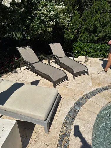 Three outdoor lounge chairs positioned on a light-colored stone patio next to a swimming pool.