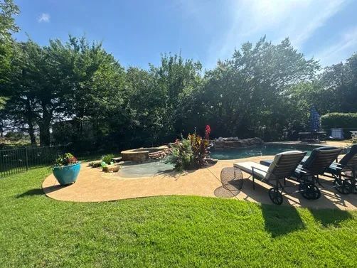A sunny backyard pool area with lounge chairs, a fire pit, and a teal planter on a textured patio surrounded by lawn.