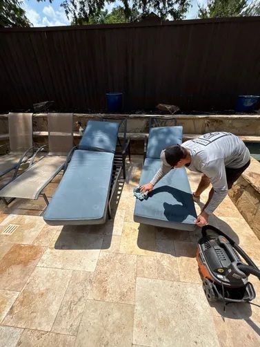 A person cleans blue outdoor lounge chairs on a stone patio with a portable vacuum nearby.