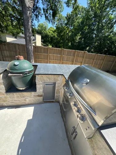 An outdoor kitchen with a green kamado grill and a stainless steel gas grill set into a stone counter by a wooden fence.