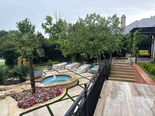 A backyard patio featuring a small circular fountain, lounge chairs, a palm tree, and stone steps leading to a house.