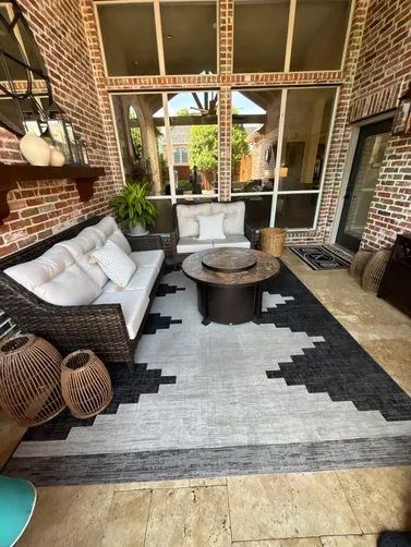 A patio with a wicker sofa, round fire pit, and black and white patterned rug against a brick wall.
