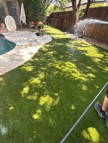 Water sprays from a hose over green artificial turf in a backyard next to a swimming pool.