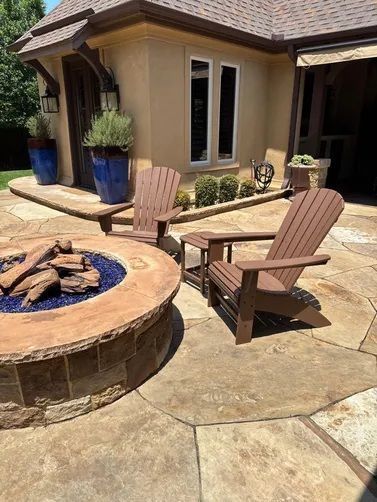 Two brown Adirondack chairs by a stone fire pit with blue glass, set on a tan patio beside a house with blue planters.