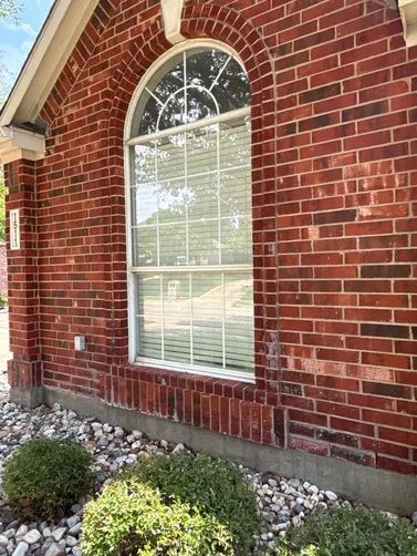 A brick home exterior with an arched window, white blinds, and decorative stone landscaping at the base.
