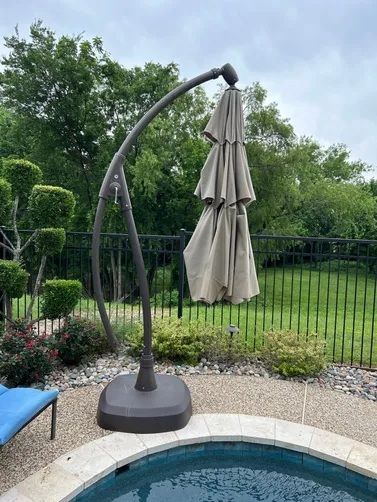 A closed, beige cantilever patio umbrella stands on a stone patio next to a swimming pool with a fence and trees behind.