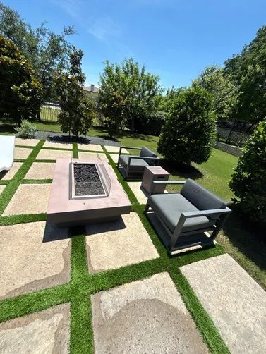 Modern outdoor patio with a rectangular fire pit, two gray lounge chairs, and square concrete pavers with grass borders.