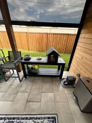 A covered patio featuring a dark wood bench with a small house-shaped bird feeder and bowls overlooking a grassy yard.