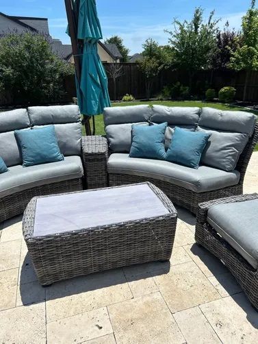 A curved grey wicker patio sectional with blue cushions and a matching coffee table on a stone patio in a sunny yard.