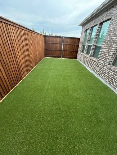 A narrow backyard featuring synthetic green turf, bordered by a wooden fence on one side and a brick house wall on the other.