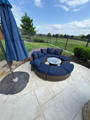 A curved, dark wicker sectional sofa with navy blue cushions and a center glass table on a backyard patio.