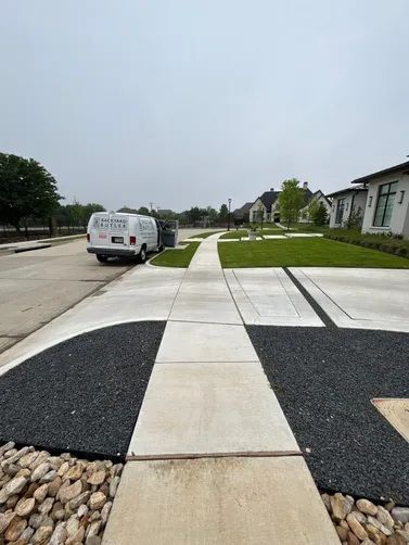 A concrete sidewalk leads toward suburban houses, flanked by dark gravel patches and a parked white van on the left.