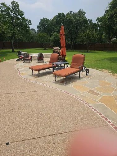 Two orange chaise lounges with a small table and umbrella on a stone patio in a backyard with green grass and trees.